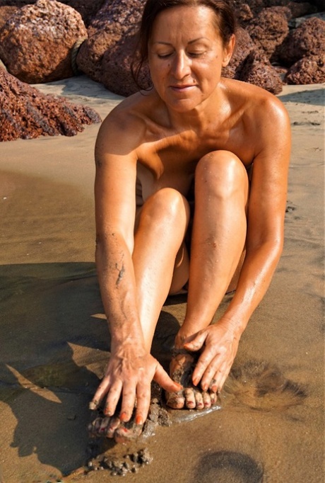 Naked older woman Diana Ananta covers her feet in beach sand at low tide