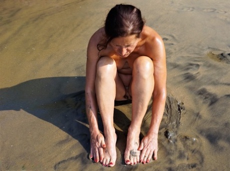 Naked older woman Diana Ananta covers her feet in beach sand at low tide